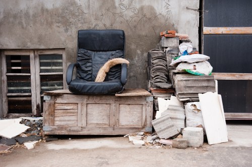 A black office chair with a padded seat, backrest, and armrests is positioned on a wooden crate situated against a weathered, grey concrete wall. The chair's fabric appears slightly worn and has a beige cushion draped over one armrest. To the right of the chair, there is a stack of building materials including concrete blocks, stone slabs, and a pile of mixed debris, which appears to be discarded waste from a construction or renovation project. Some of the blocks are stacked directly on the ground, while others rest on top of a makeshift pile. Scattered debris, such as broken pieces of concrete and small fragments, are visible on the pavement around the pile. The background includes a vent opening at ground level and a partially visible black roller shutter door to the far right. The scene is outdoors, set on a concrete or tarmac surface, suggestive of a service yard or back alley typical of commercial waste collection areas in Upminster, indicating it is related to the rubbish removal services offered by Commercial Waste Upminster.