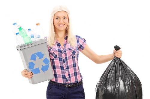 A young woman with blonde hair, wearing a casual pink and blue checkered shirt and dark trousers, standing against a plain white background, holding a small grey recycling bin with a blue recycling symbol in her left hand filled with clear plastic bottles of various sizes and colours, and a full black rubbish bag in her right hand. The scene appears to be a staged presentation or educational image related to waste disposal or recycling practices, with no environmental context visible. The woman's expression is neutral and friendly, and the focus is on her holding the waste containers, emphasizing waste management actions in general. This image could be associated with waste removal services in Upminster or similar areas, illustrating responsible rubbish handling or recycling efforts, without contextual clutter or environment details.