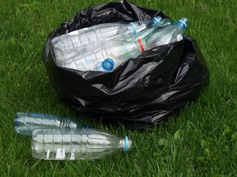 A black plastic rubbish bag placed on a grassy outdoor area, containing several clear plastic water bottles with blue caps, some of which are partially visible through the open top of the bag. Two additional bottles are lying on the grass, one near the bag and another further away. The scene appears to be outdoors, with green grass and some blades of grass visible in the foreground and background, suggesting a natural or garden setting. The bottles are transparent, with textured ridges on their bodies, and the overall lighting is natural, highlighting the textures of the plastic and the vibrant green of the grass. This image relates to rubbish disposal and waste collection services, such as those provided by Commercial Waste Upminster, in the context of managing waste in the local area near the postcode in Upminster.