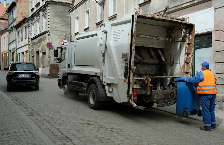A row of large waste bins with green, red, blue, and dark green lids are positioned along a paved pathway beside a brick wall. The bins are made of durable plastic with textured surfaces on the lids a