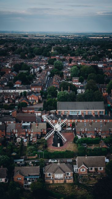 An aerial view of a residential area featuring a traditional windmill situated amongst rows of brick houses and terraced homes. The windmill has white blades and a dark, conical roof, positioned centr