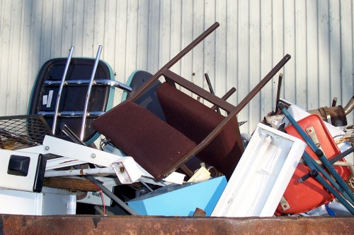 A large pile of assorted discarded items and waste materials stacked outdoors against a corrugated metal wall. The collection includes a black plastic chair with metal legs, a brown upholstered armchair with wooden armrests, and a white plastic frame, possibly from a broken appliance or furniture piece. Also visible are a blue plastic storage container and various other plastic and metal objects, including a red and blue trolley or cart. The items appear to be weathered and randomly piled, indicating collection for rubbish removal, with some objects leaning or resting against each other. The scene suggests a typical outdoor waste collection area in Upminster, with natural light highlighting the textures and colors of the mixed refuse, reflecting the type of clutter that Commercial Waste Upminster often manages for local businesses or residents undergoing clearance or disposal operations.